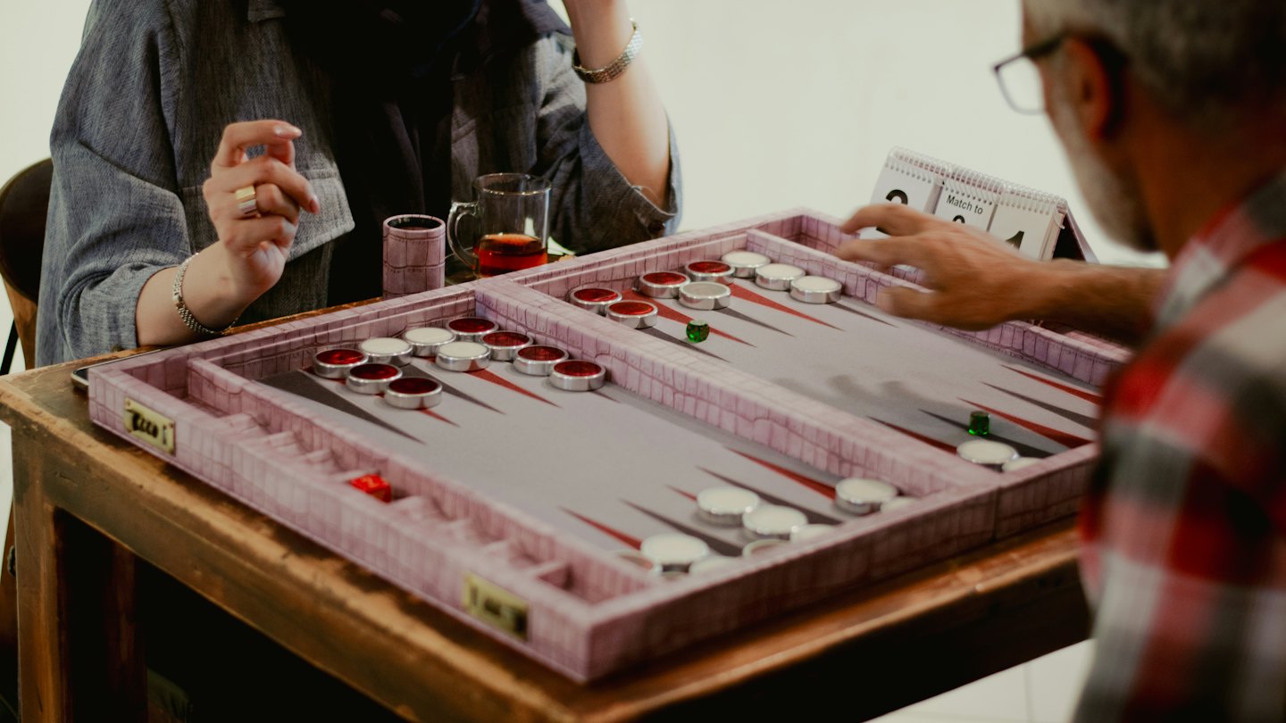 Two people play a game of backgammon on a wooden table.
