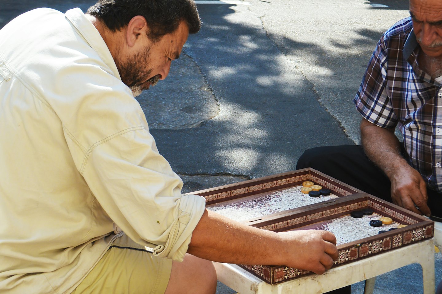 Two men play a game of backgammon on an outdoor stool.